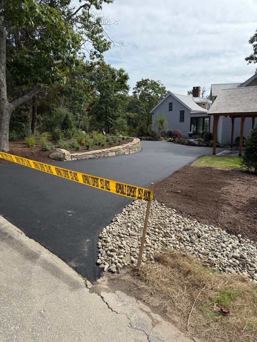 Freshly paved asphalt driveway with yellow caution tape, leading to a house with landscaping and trees.