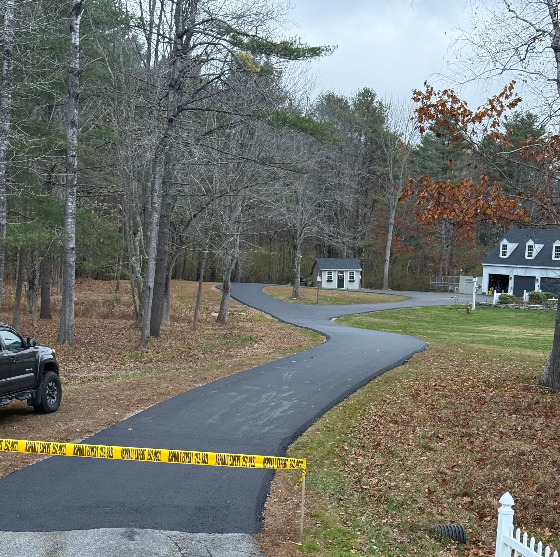 Winding driveway through leafless woods, with caution tape in front and houses in the background.