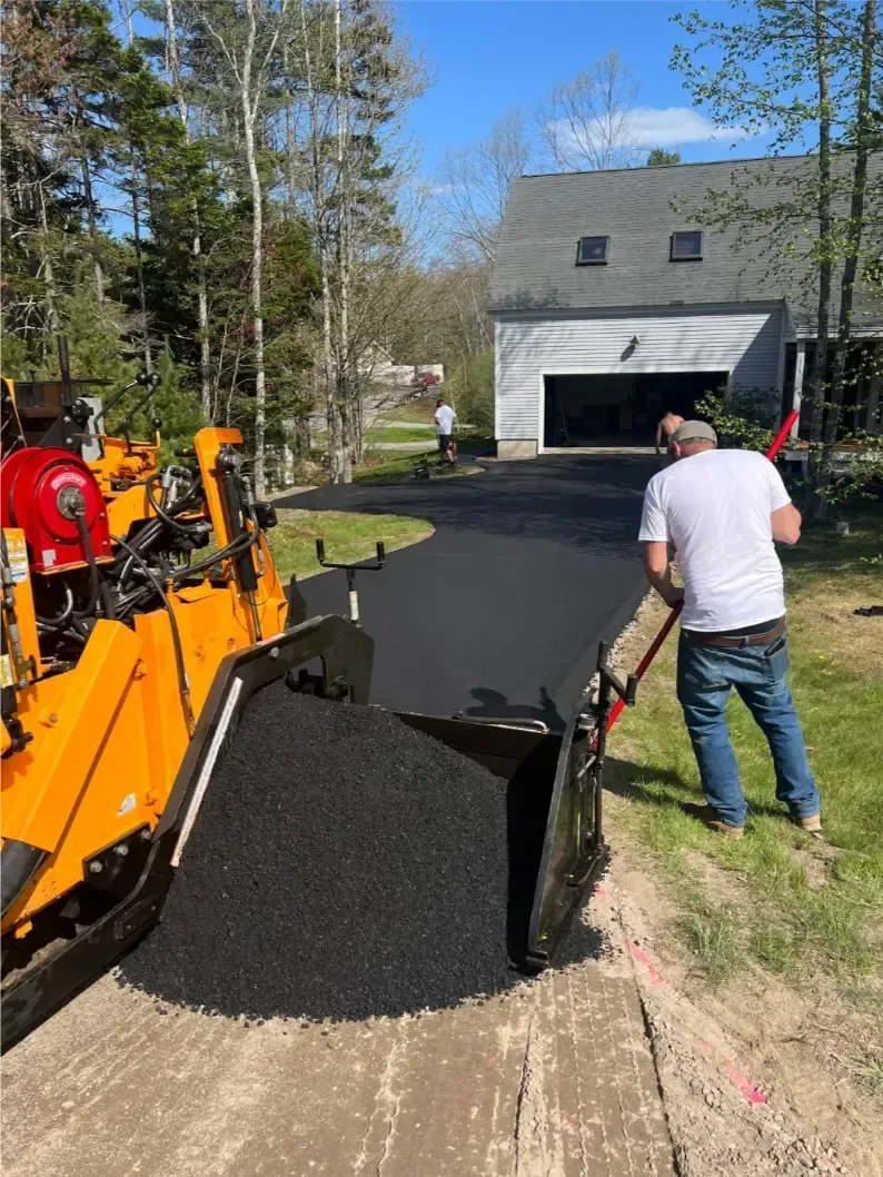 Blacktop being poured from an orange paver by a driveway beside a gray garage.