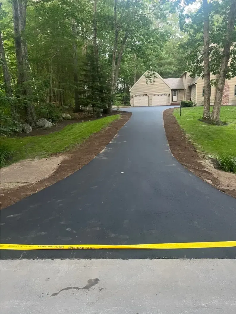 Newly paved black asphalt driveway leading to a house with a two-car garage. Yellow caution tape across the bottom.