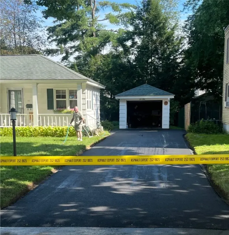 A residential driveway blocked by yellow caution tape. Houses and a garage are visible. A person is working in the yard.