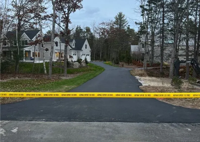 Paved driveway with yellow caution tape, leading to houses in a wooded area.