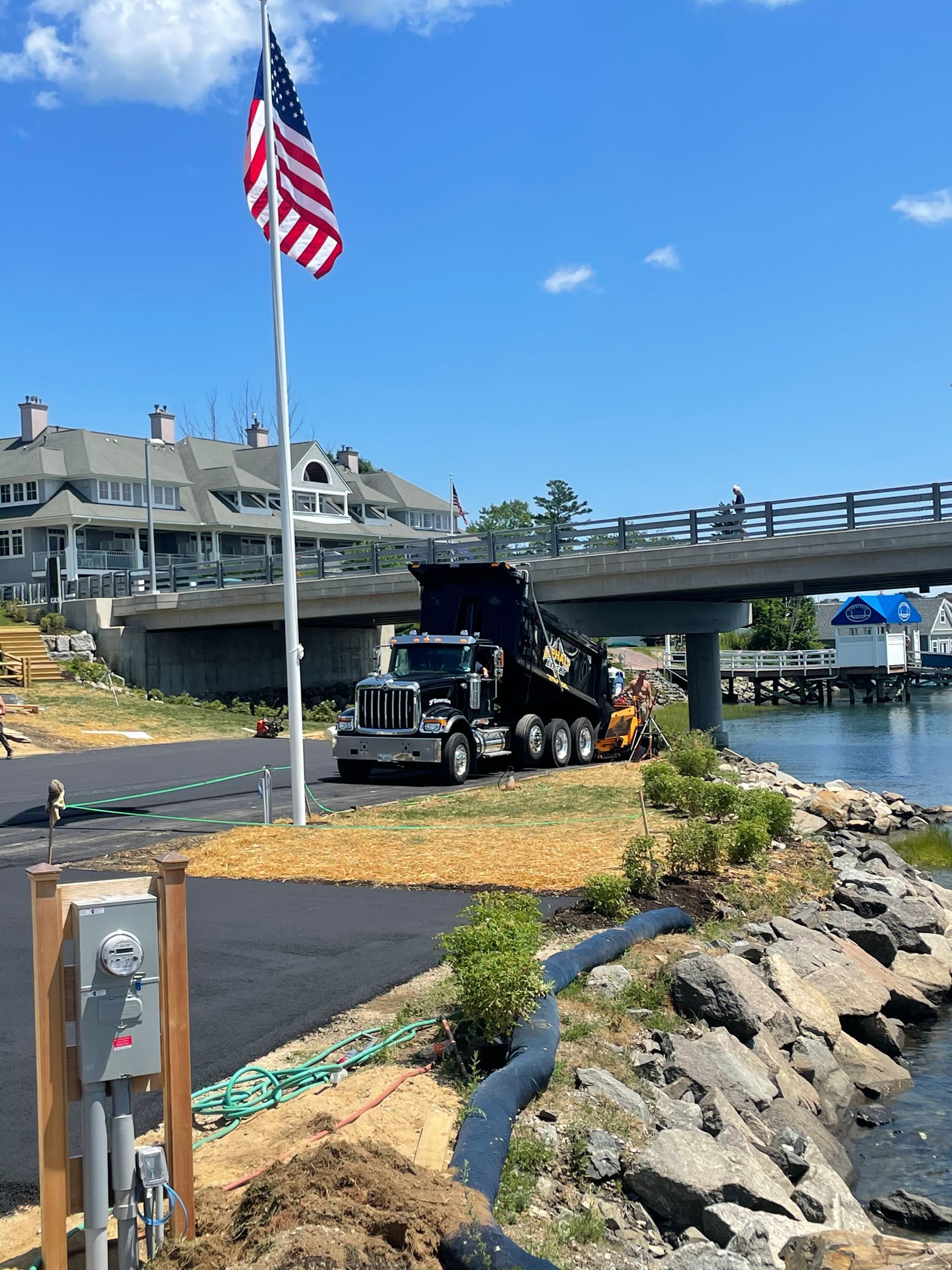 A black dump truck unloading gravel near a bridge and water under a blue sky, an American flag waves.