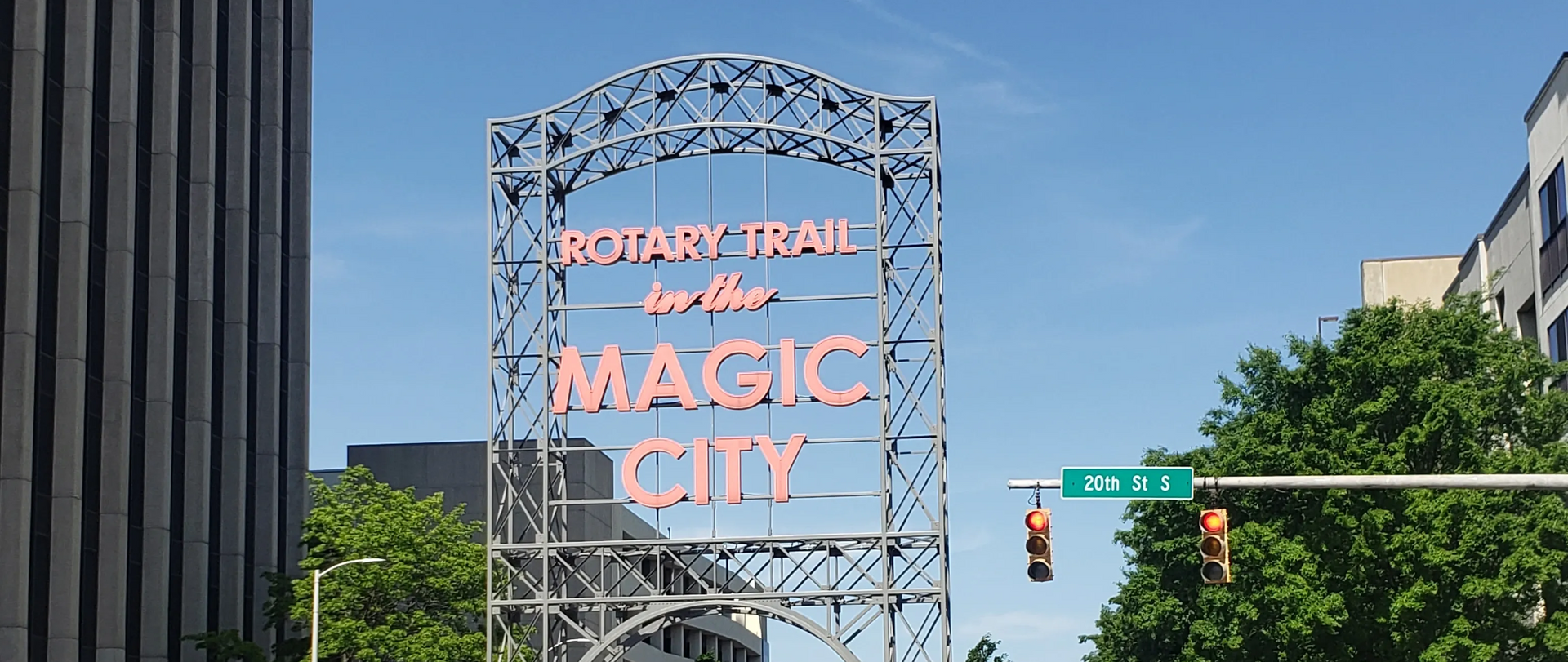 Neon “Maggie City” sign on a downtown street under a clear blue sky, with buildings and traffic light nearby