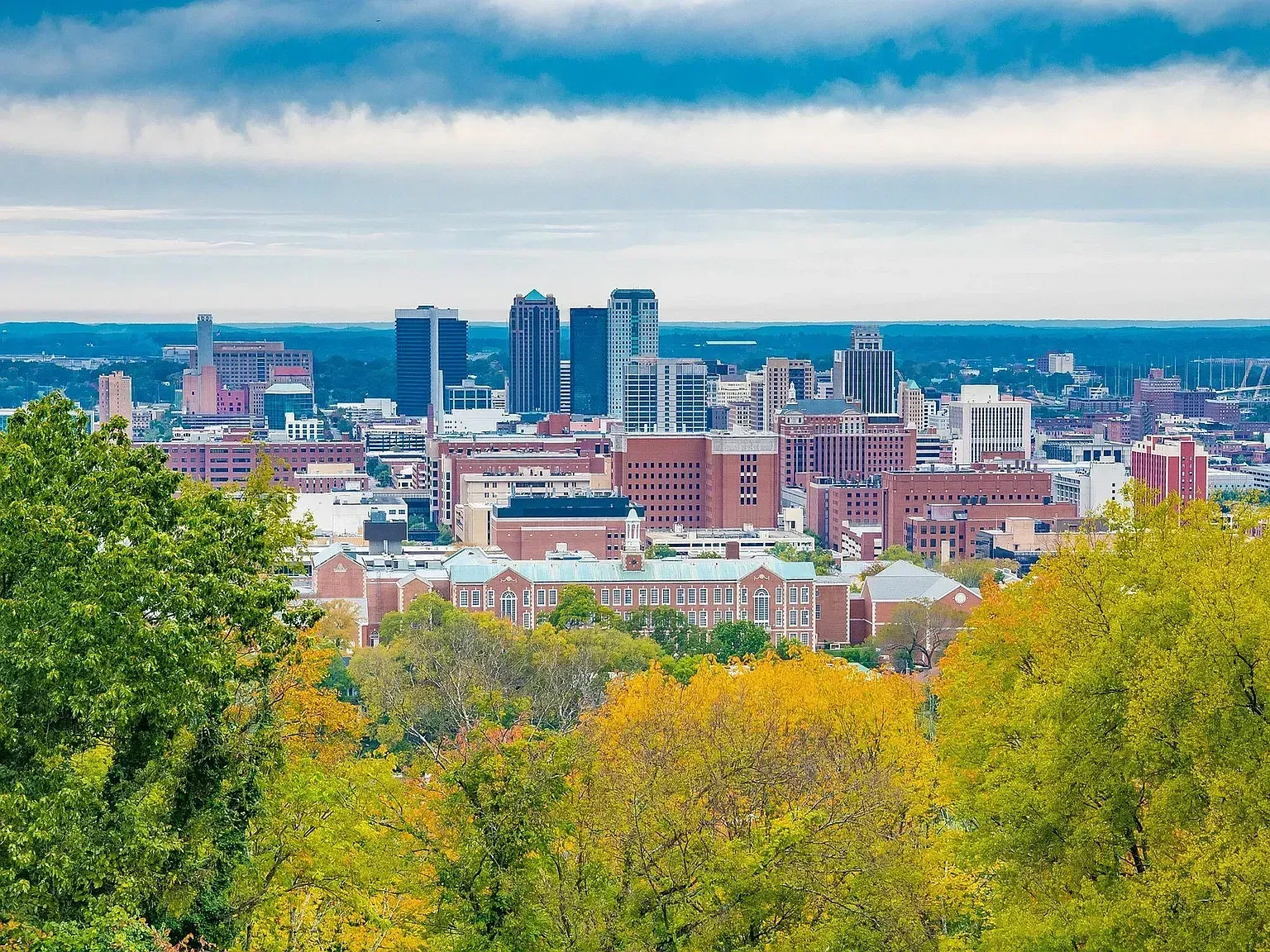 City skyline with modern buildings above colorful autumn trees under a cloudy blue sky