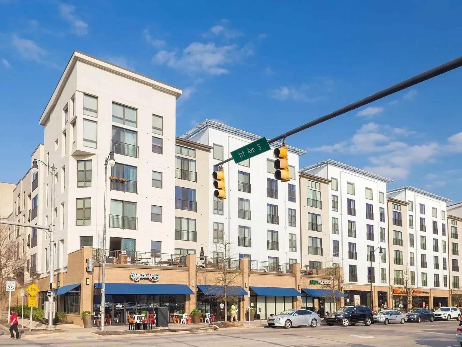 Street corner with white apartment buildings, storefronts, cars, and traffic lights under a blue sky