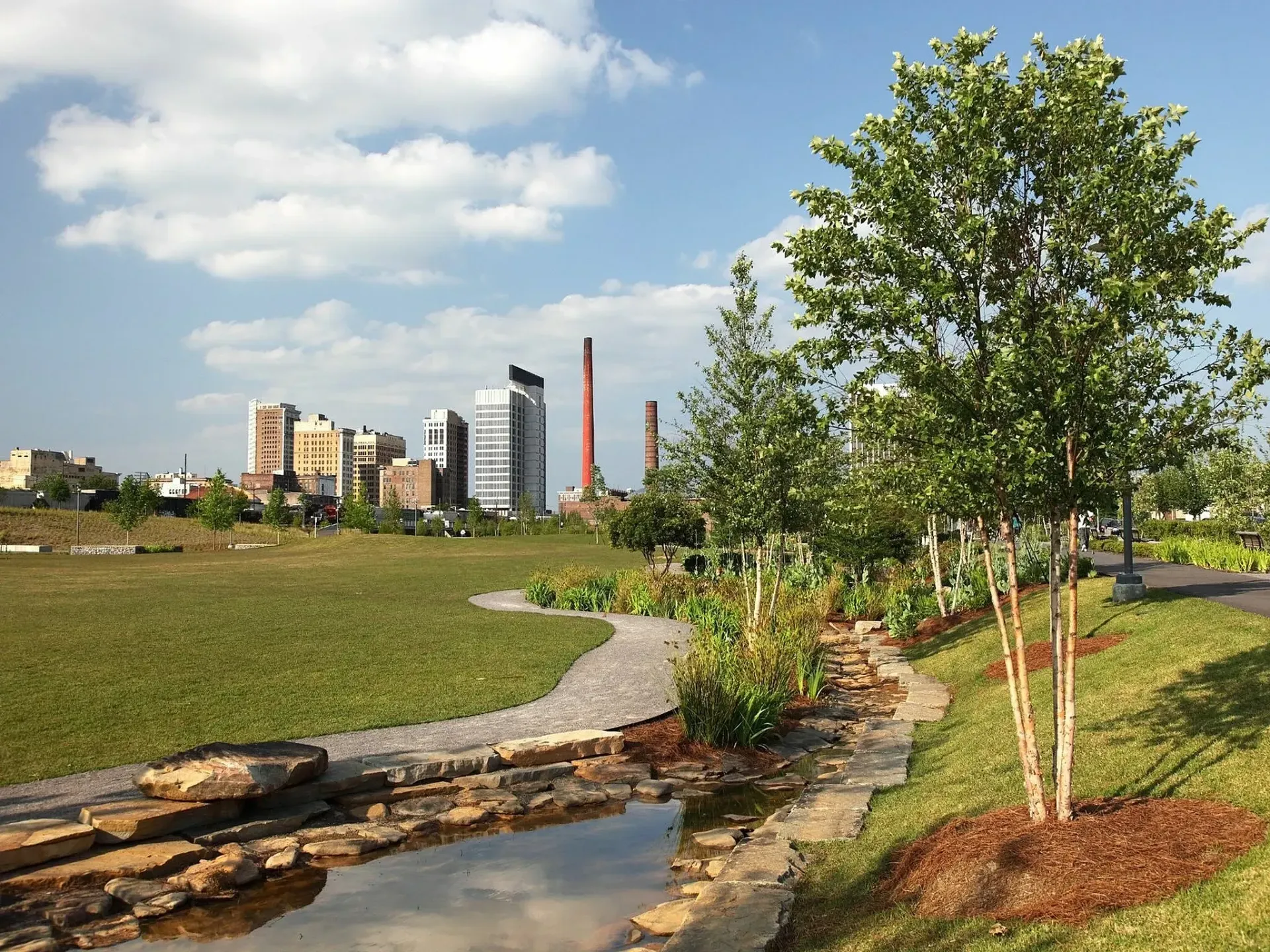 Park with pond, walking path, trees, and city skyline under a blue sky with clouds