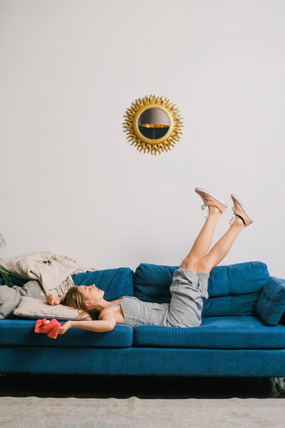 Person lying on a blue couch with legs raised in a white living room, under a small wall mirror.