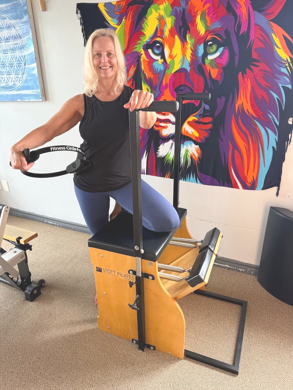 Woman exercising on a Pilates chair beside a colorful lion mural in a gym.