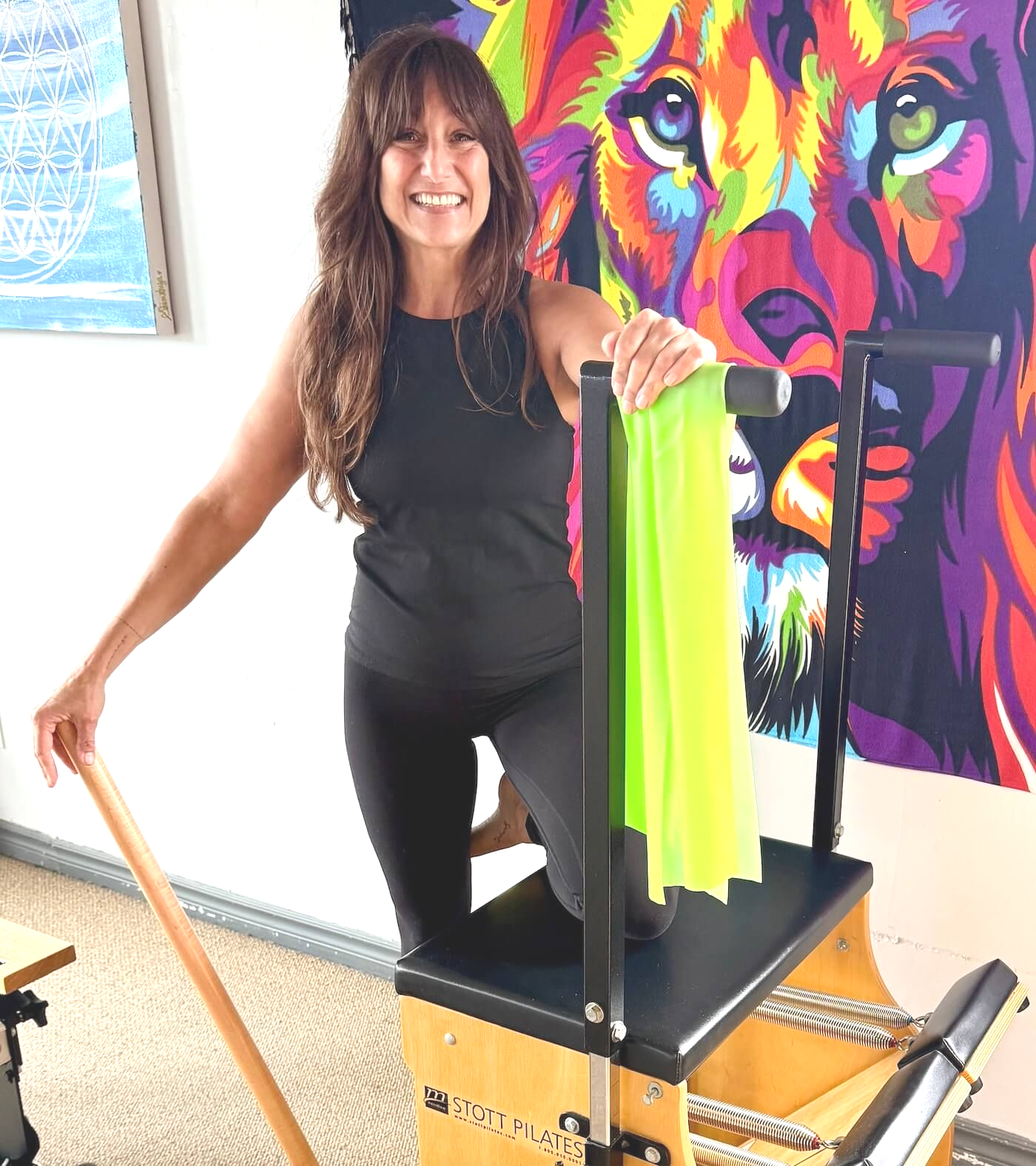Woman standing beside a Pilates chair with neon straps, smiling in front of a colorful lion painting