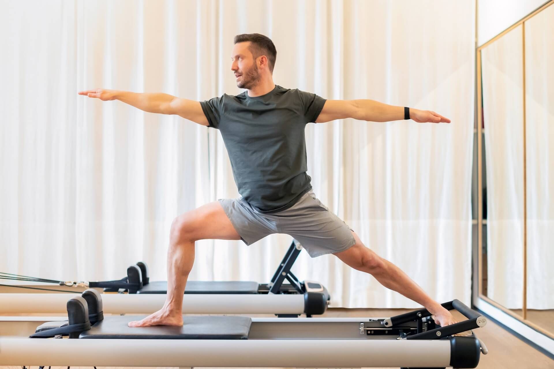 Man balancing in Warrior II pose on a Pilates reformer in a bright studio