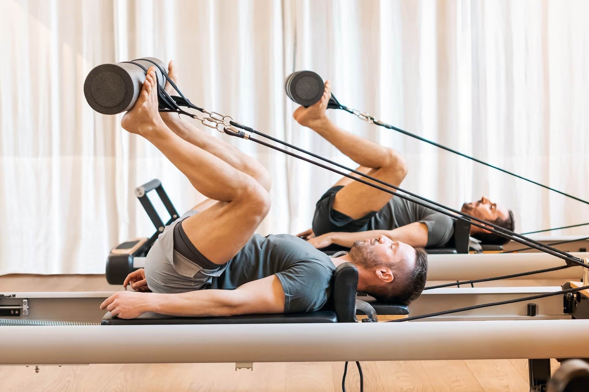 Two men doing resistance band leg exercises on reformer beds in a bright studio