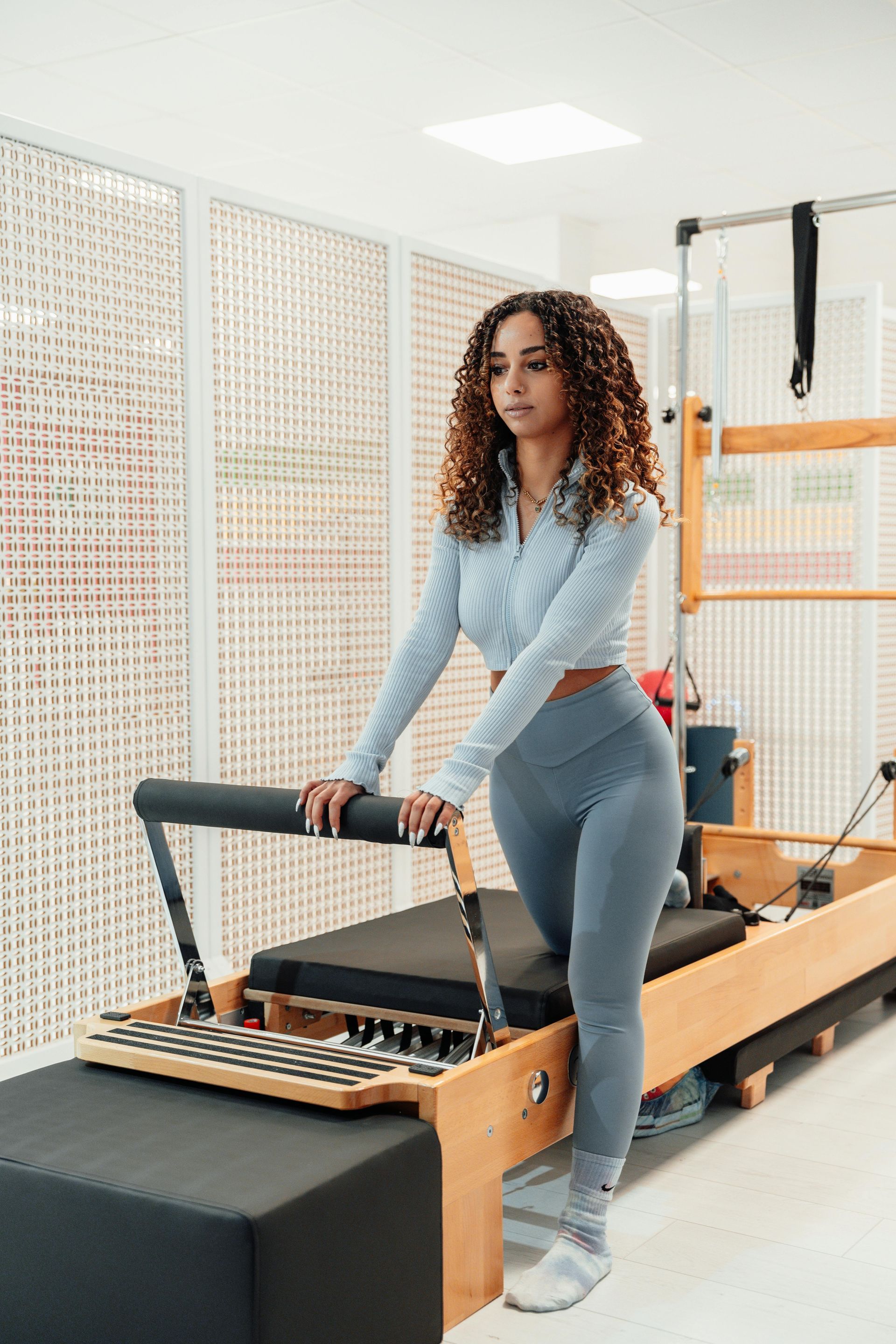 Woman using a Pilates reformer in a bright studio, wearing gray workout clothes.