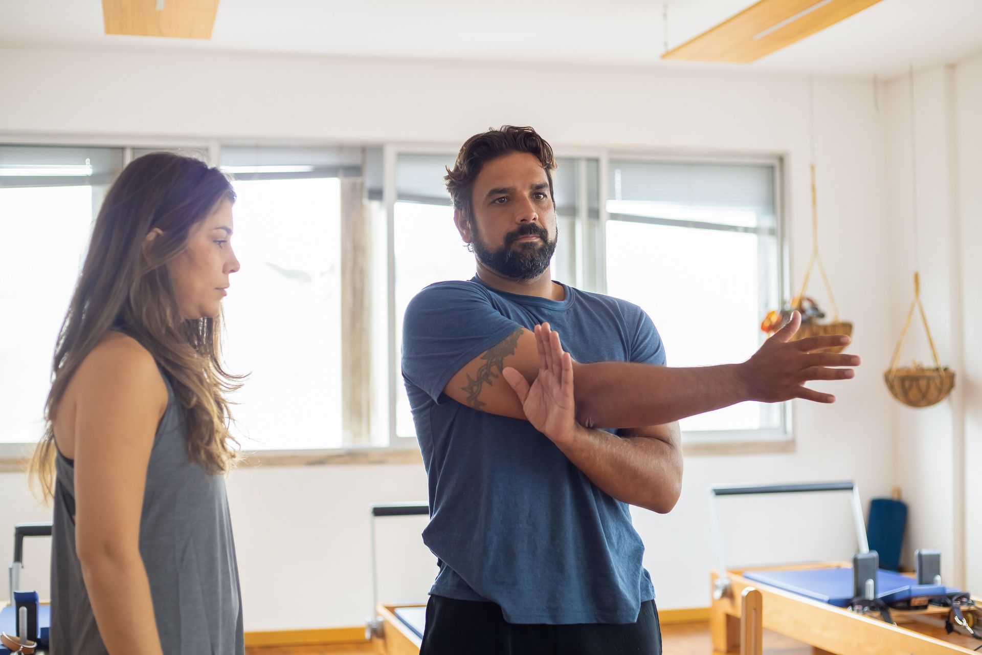 Two people in a bright room, one demonstrating an arm stretch while the other watches.
