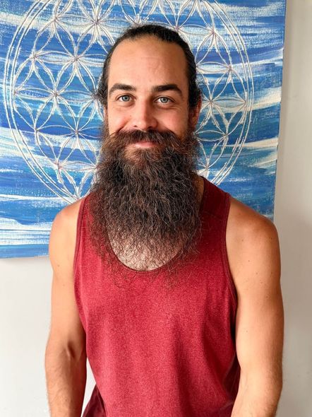 Smiling person with long beard in red tank top, standing before a blue patterned tapestry