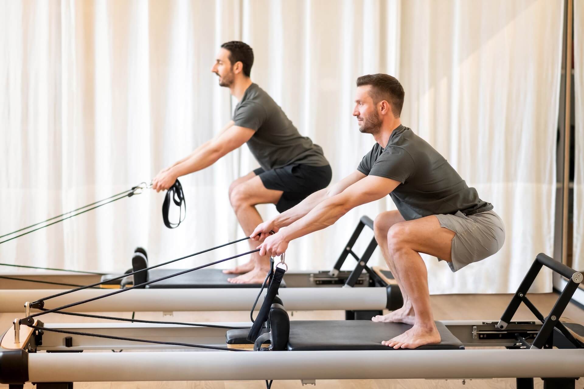 Two men exercising on Pilates reformers in a bright studio, pulling straps in a squat position