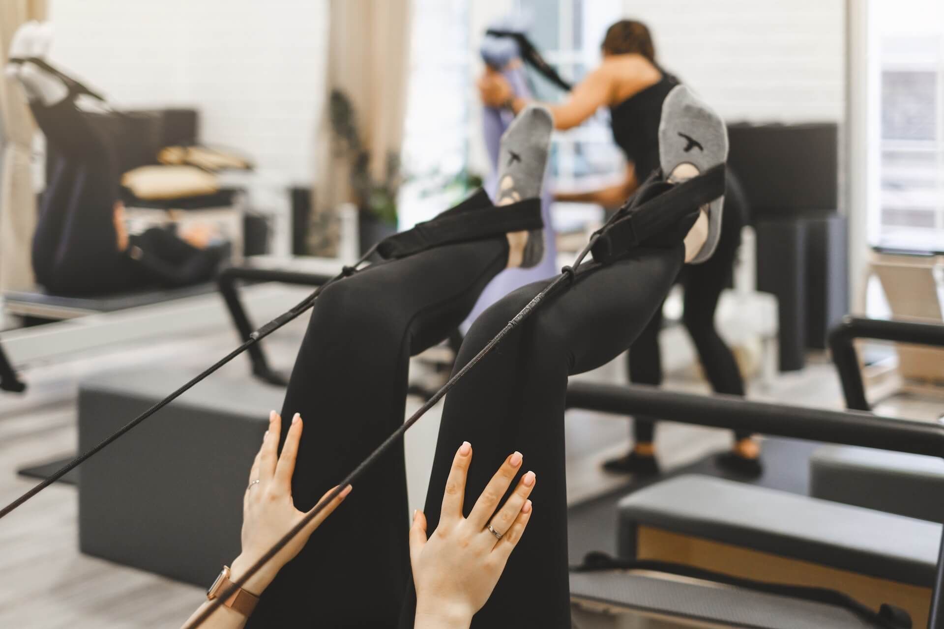 Person exercising on Pilates apparatus with feet in black straps, wearing black leggings.