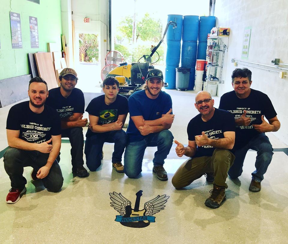 Six people kneeling in a bright room, smiling and posing together in front of blue barrels.