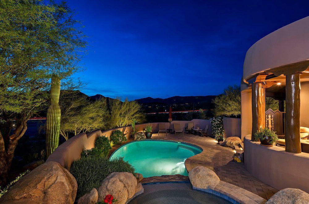 Outdoor swimming pool at dusk with mountain views, surrounded by rocks and a curved wall.