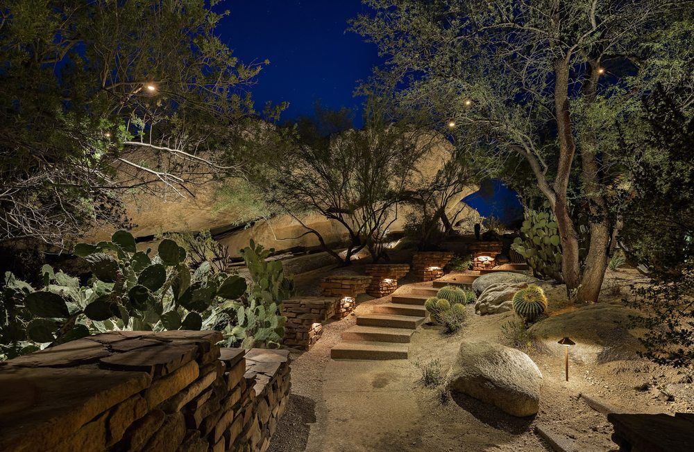 A stone pathway winds through a desert landscape at night, illuminated by soft ground lights amidst cacti and boulders.