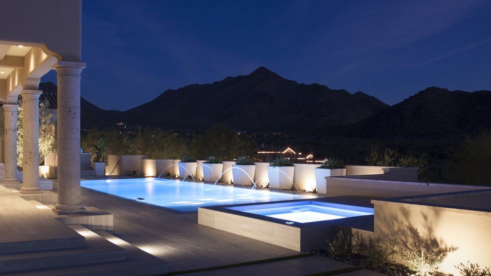 A lighted swimming pool and hot tub on a patio at dusk, set against a dark mountain landscape.