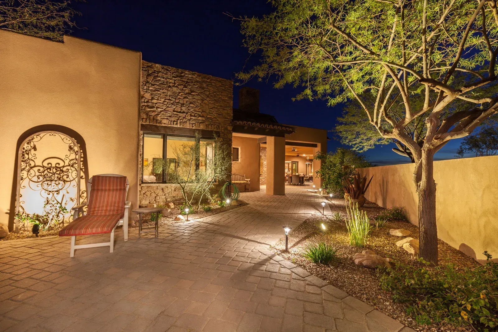 A lit stone patio at night features a metal gate, lounge chair, tree, and landscaped path leading to a stucco home.