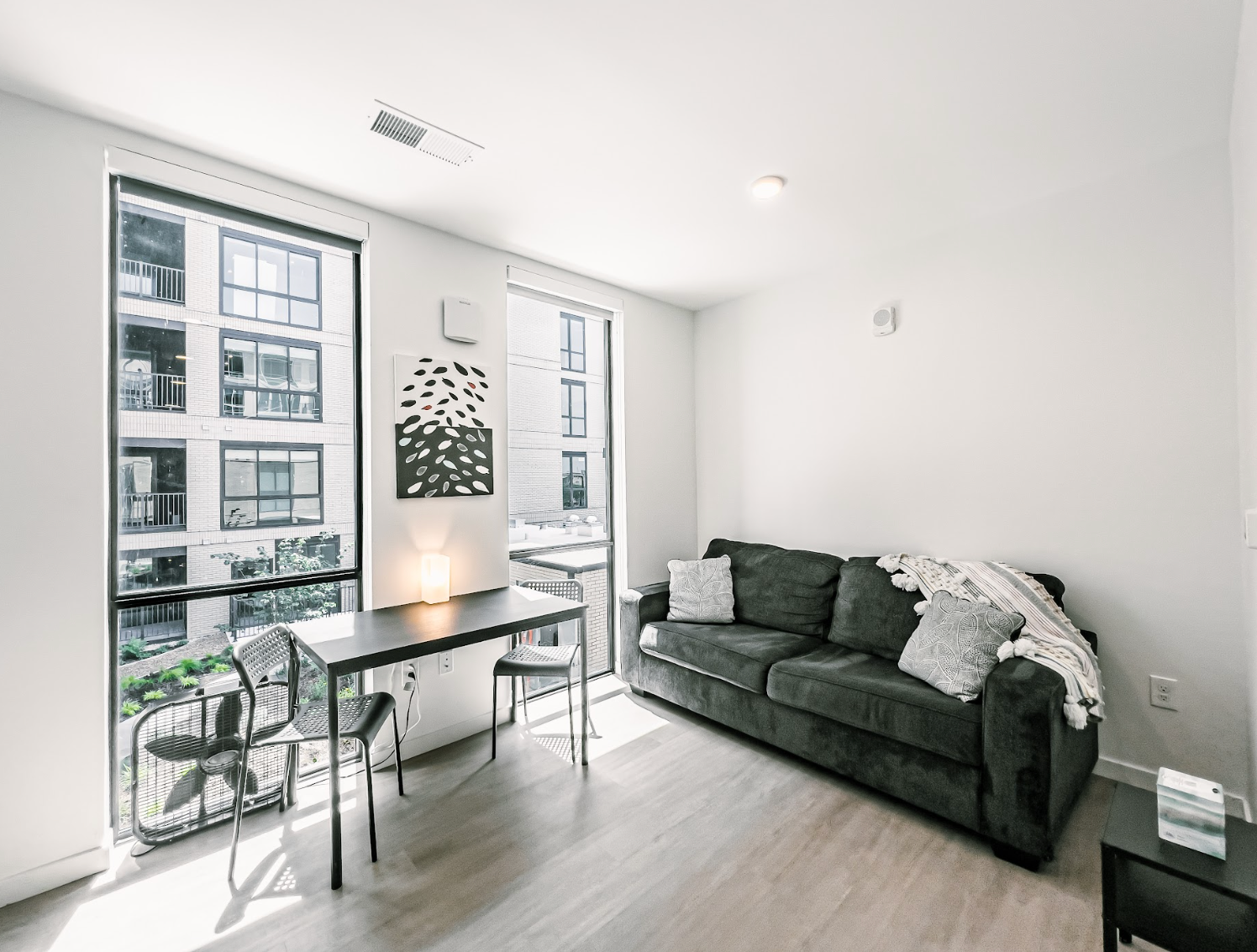 A bright living room features a dark gray sofa, a small black dining table with chairs, and a large window.