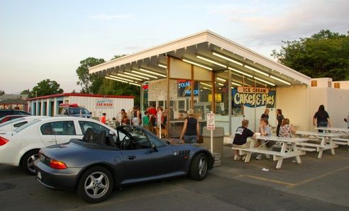 A car is parked in front of a restaurant with people sitting at picnic tables.