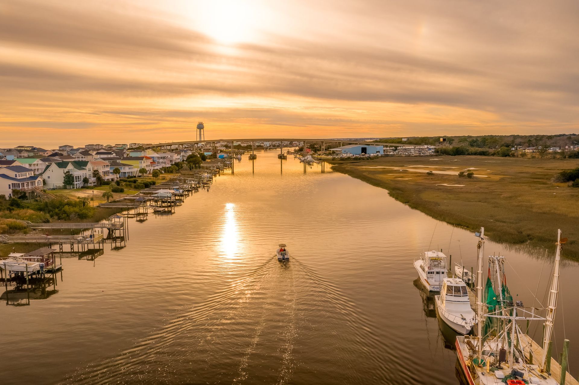 Sunset over a wide river lined with boats and houses, with a small canoe near the center and a bridge in the distance