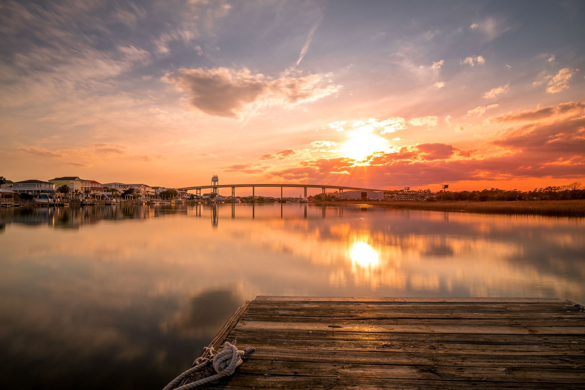 Sunset over a calm harbor with a wooden dock and bridge reflected in still water
