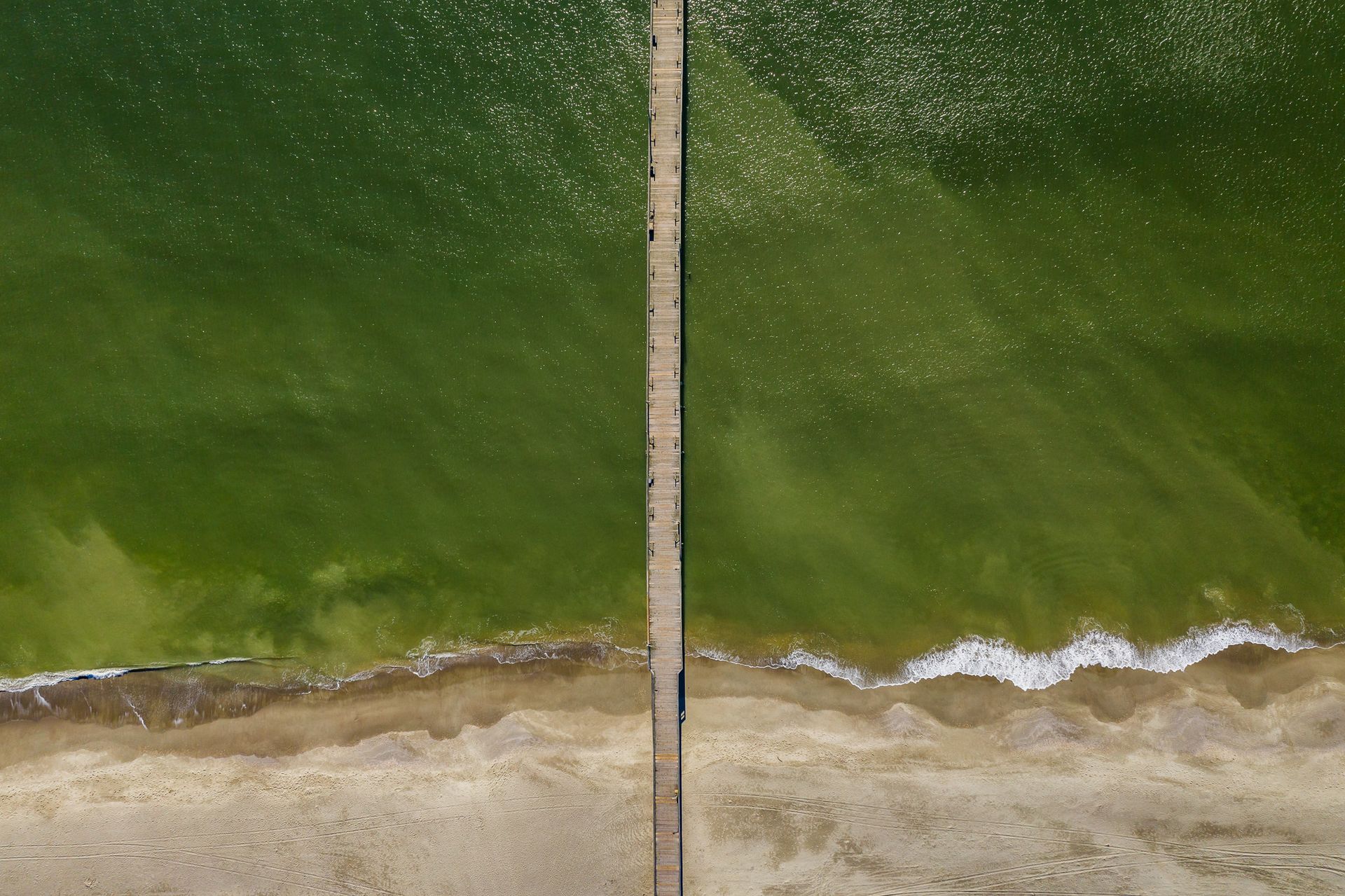 Aerial view of a green shoreline divided by a long narrow pier, with waves along the sandy beach.