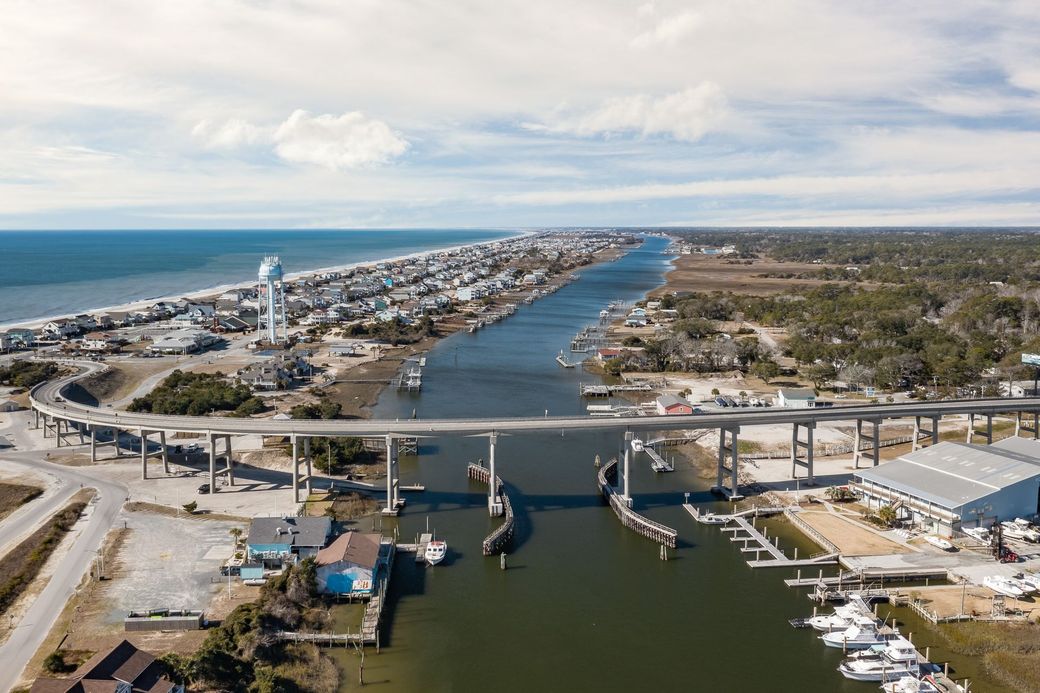 Aerial view of a coastal canal town with a bridge, boats, and ocean shoreline.