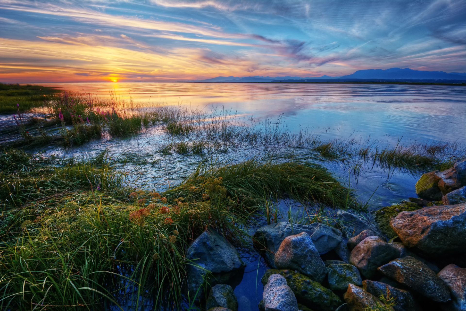 Sunset over a rocky lakeshore with grasses and calm water reflecting orange and blue sky