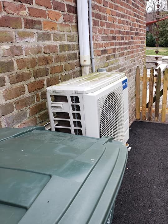 A white air conditioner is sitting on the side of a brick building next to a green trash can.