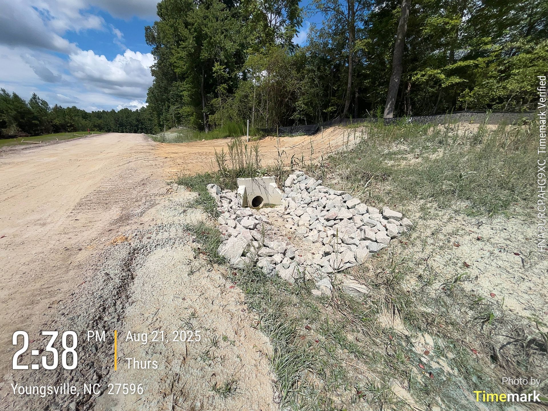 Dirt road with a culvert surrounded by rocks and weeds. Forest in the background.