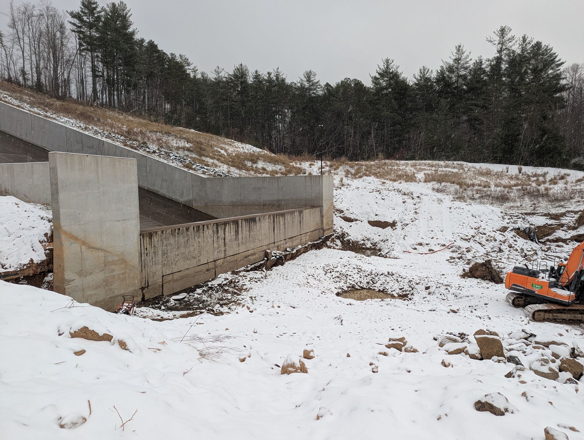 Concrete retaining wall on a snow-covered hillside with an excavator on the right and trees in the background.