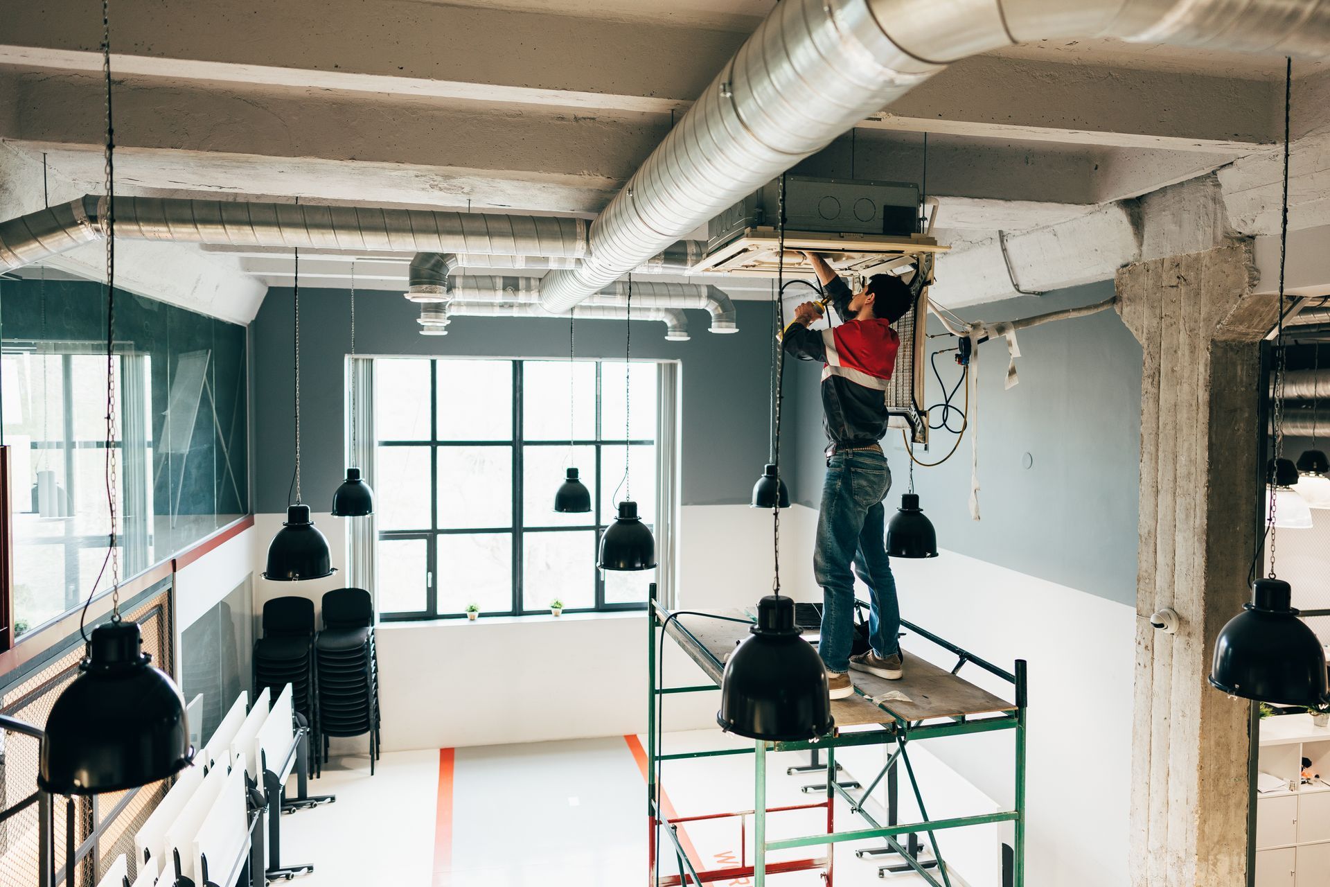 Person on scaffolding installing electrical fixture in open-plan room with exposed ductwork and pendant lights.