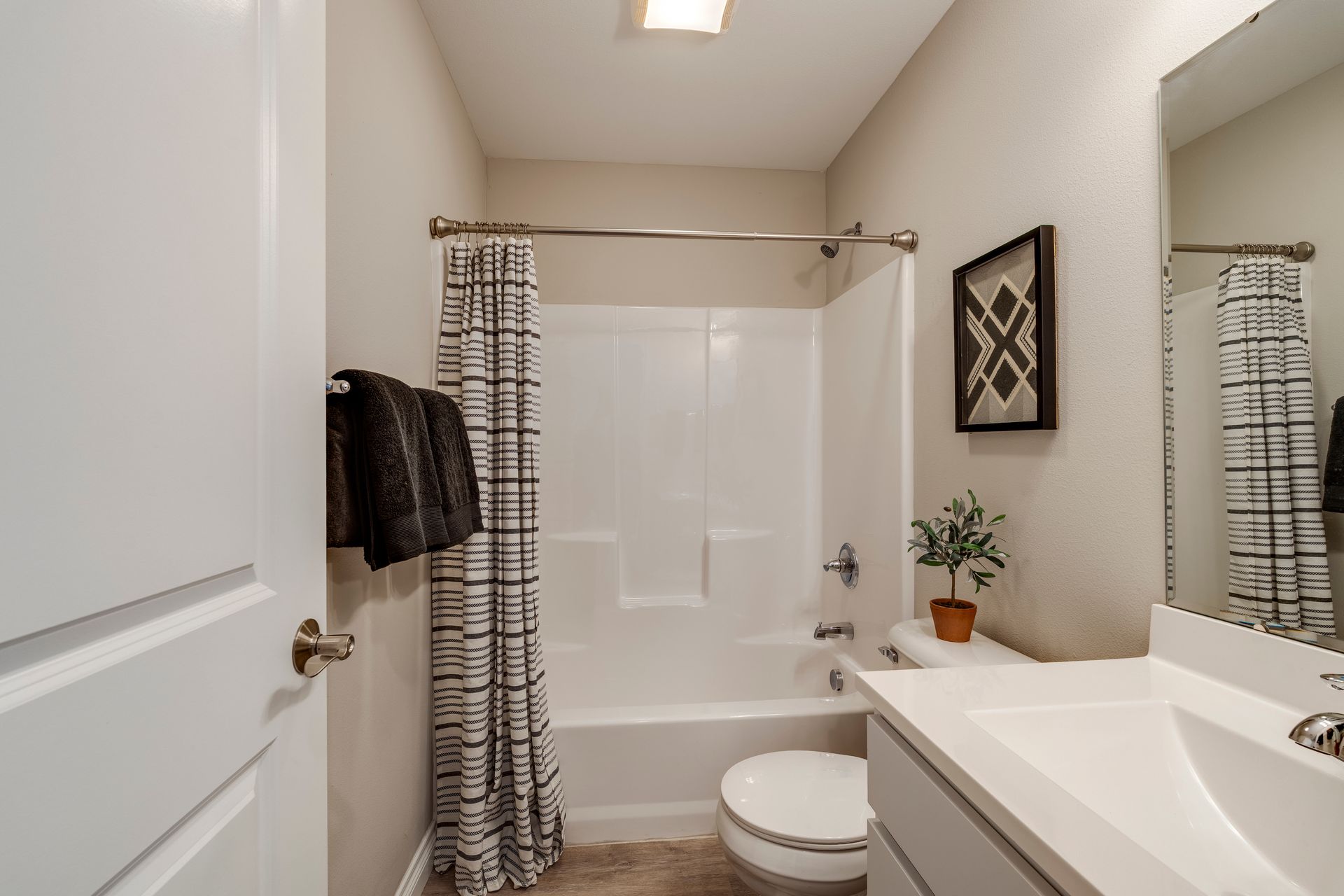 Bathroom with white walls, tub, toilet, and vanity. Black and white shower curtain, small plant, and framed artwork.