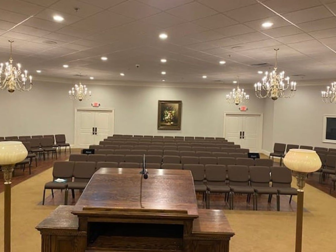 Empty auditorium with rows of brown chairs, a wooden lectern, chandeliers, and doors.