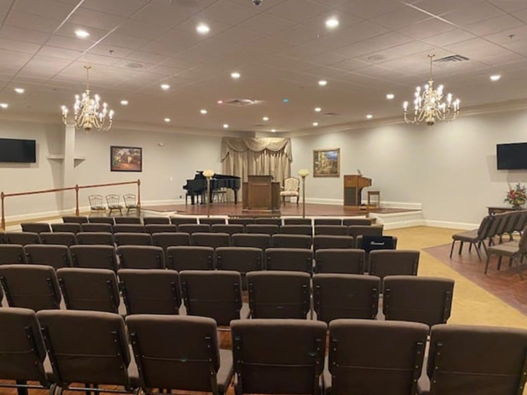 Rows of chairs face a stage with a piano, podium, and draping, in a formal room.