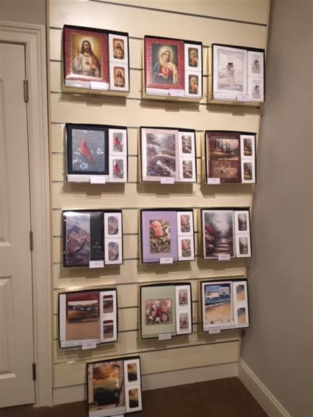 Wall display of framed art, mostly religious, with cream-colored horizontal slats.