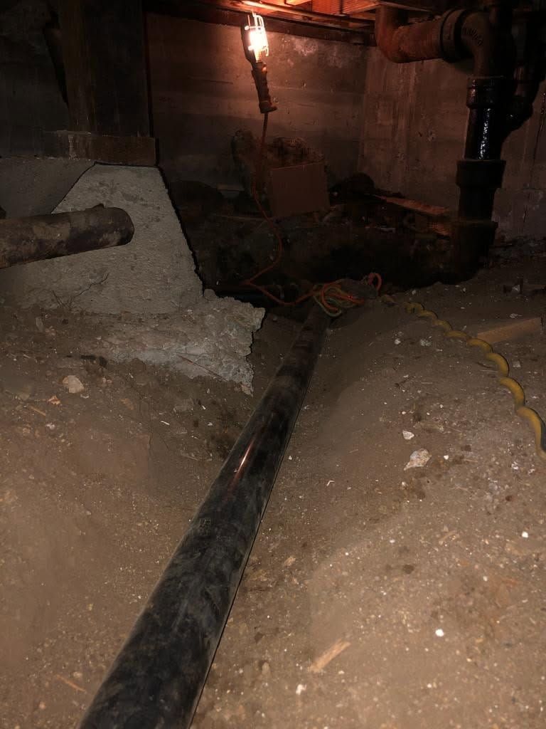 Dark basement crawlspace with a black pipe running over dirt and concrete under warm light