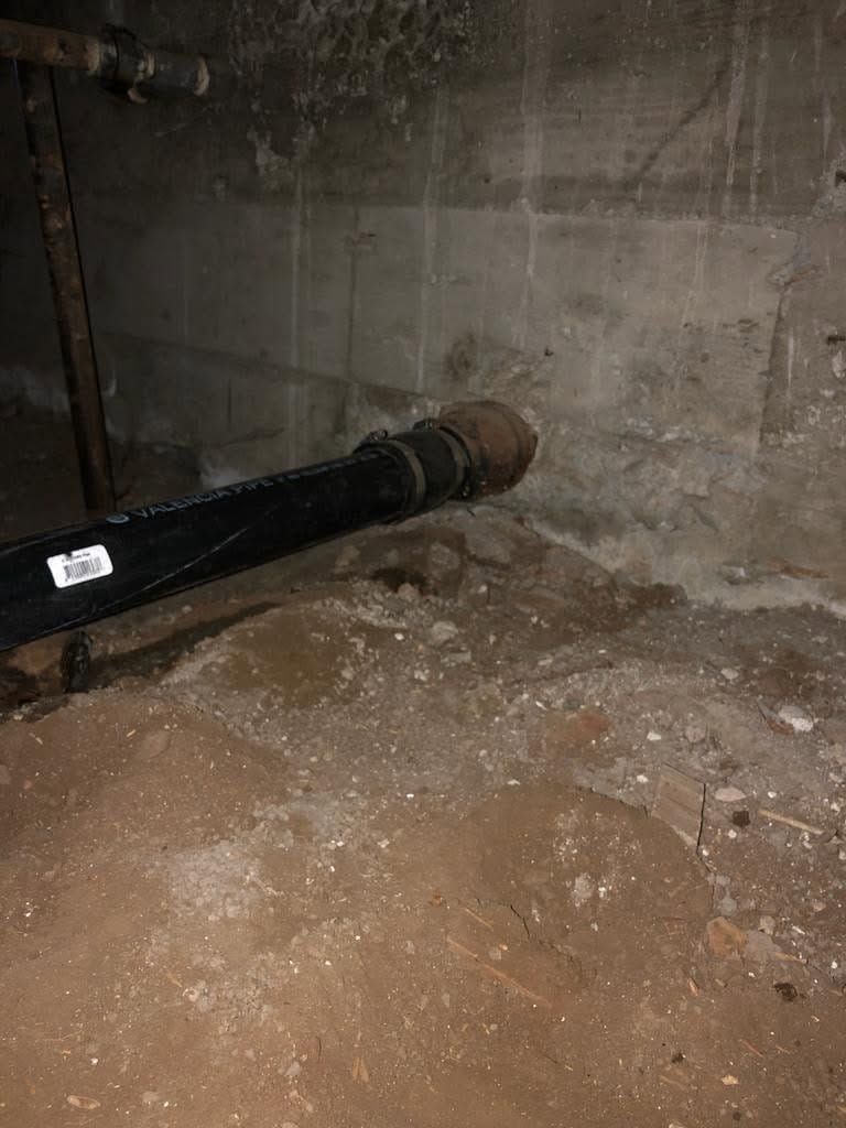 Black pipe entering a concrete wall in a dusty crawlspace or basement corner.