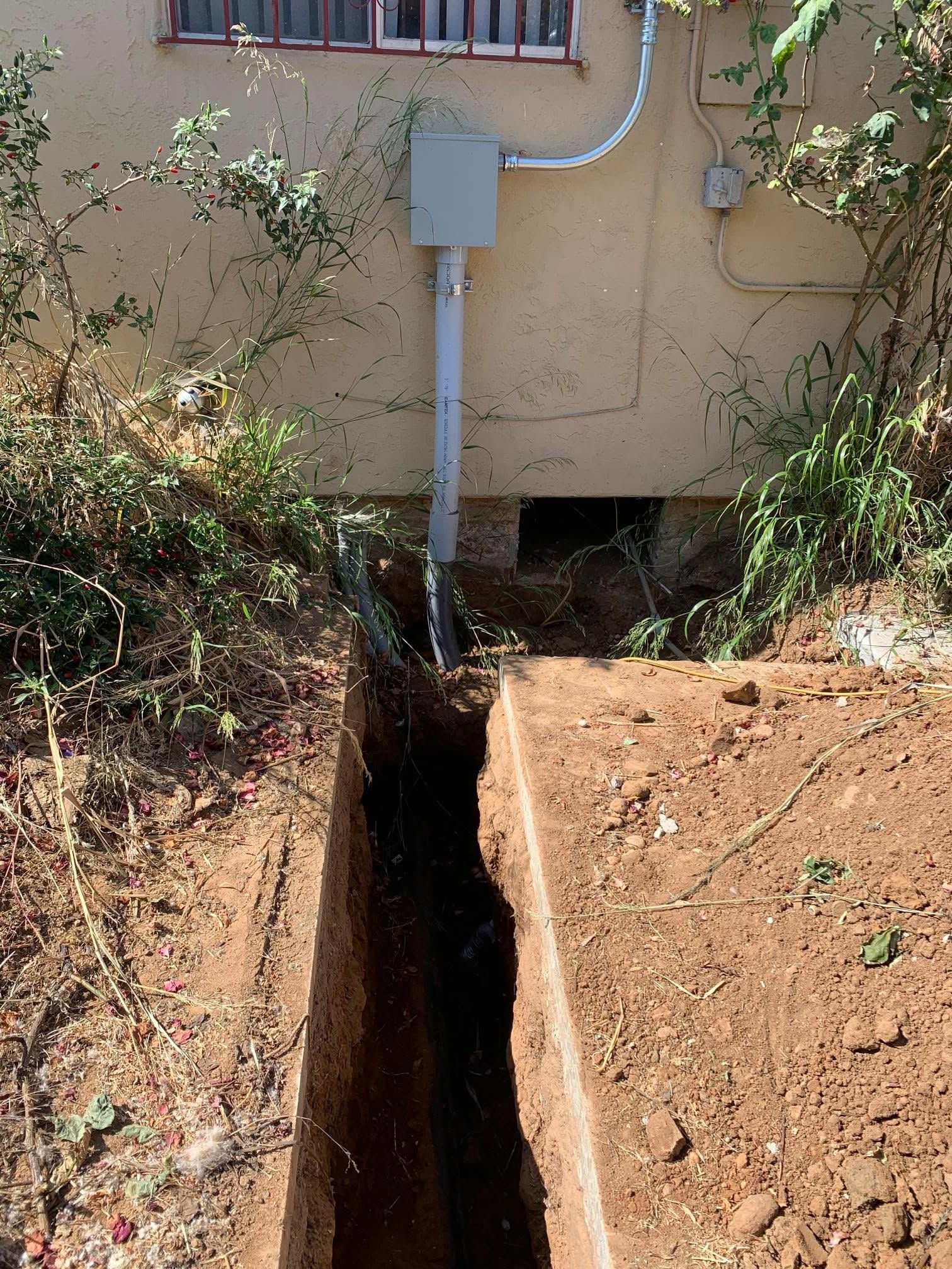 Narrow trench dug beside a house, with exposed soil and a utility conduit/electrical box above it.