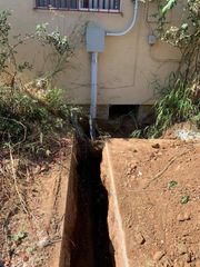 Narrow trench dug beside a house, with exposed soil and a utility conduit/electrical box above it.
