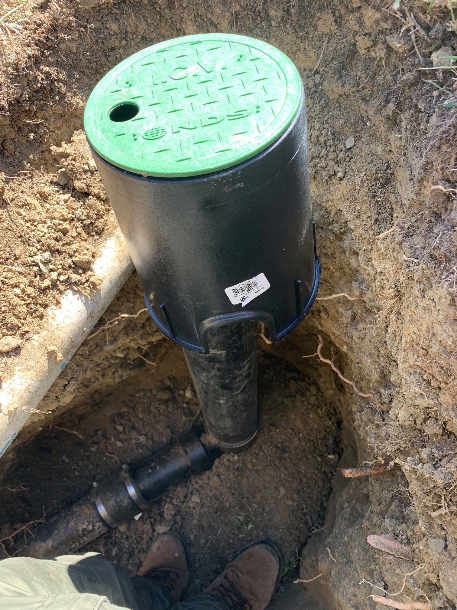 Black utility valve box with green perforated lid installed in a dirt trench with exposed pipes