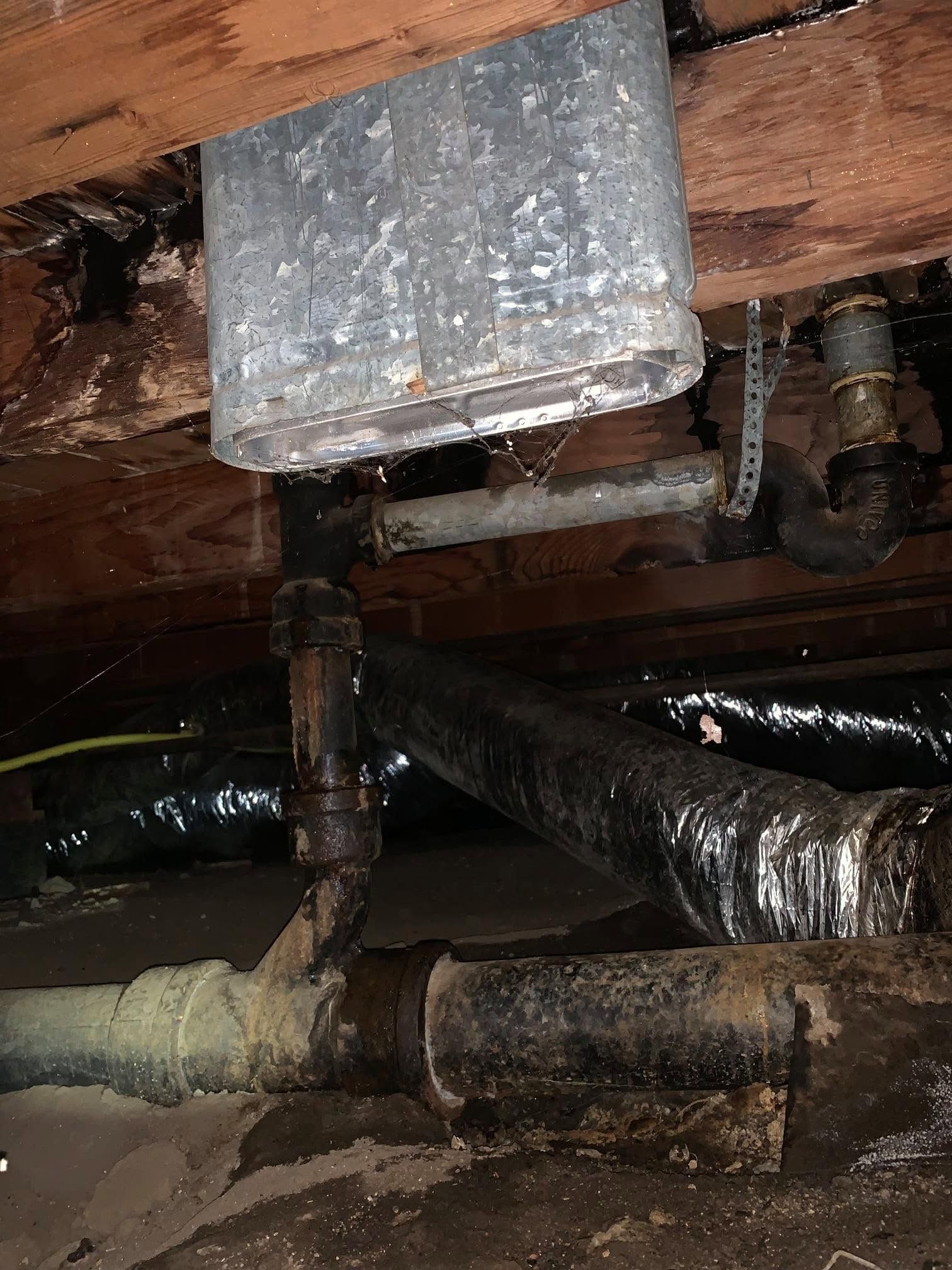 Old metal HVAC ductwork and pipes under wooden floor joists, showing rust and insulation.