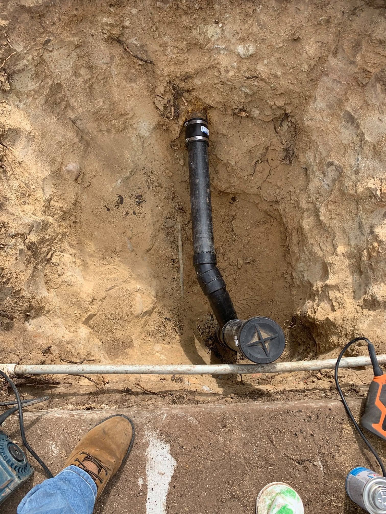 Excavated dirt trench with black pipe, valve, and construction tools beside a booted worker’s foot