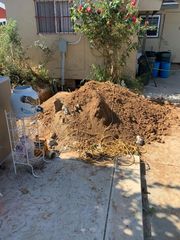 Large pile of dirt and debris in a sunny backyard beside a house and potted plants
