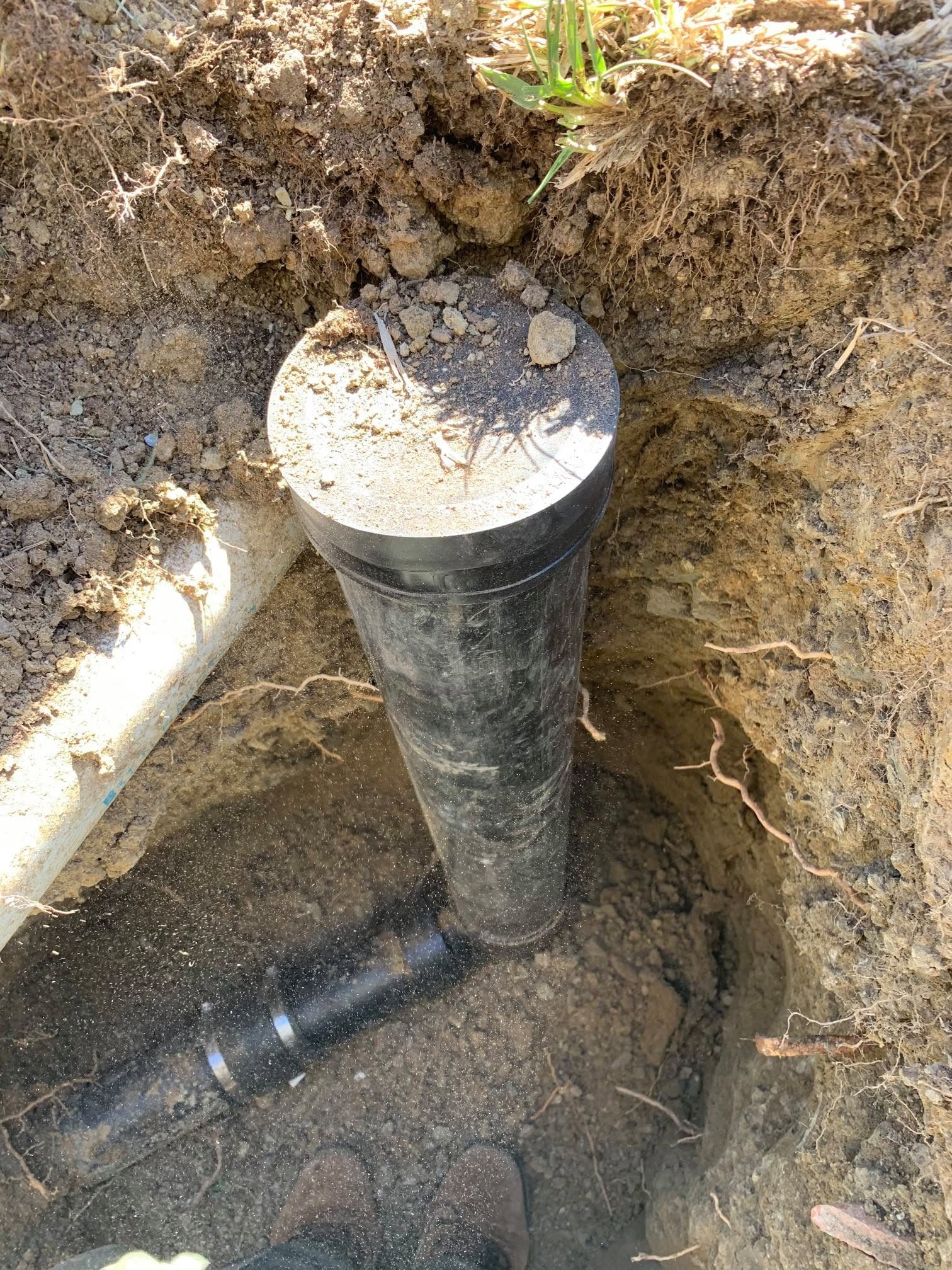 Exposed black drain pipe in a dirt trench beneath a concrete cap, with a horizontal pipe at the base.